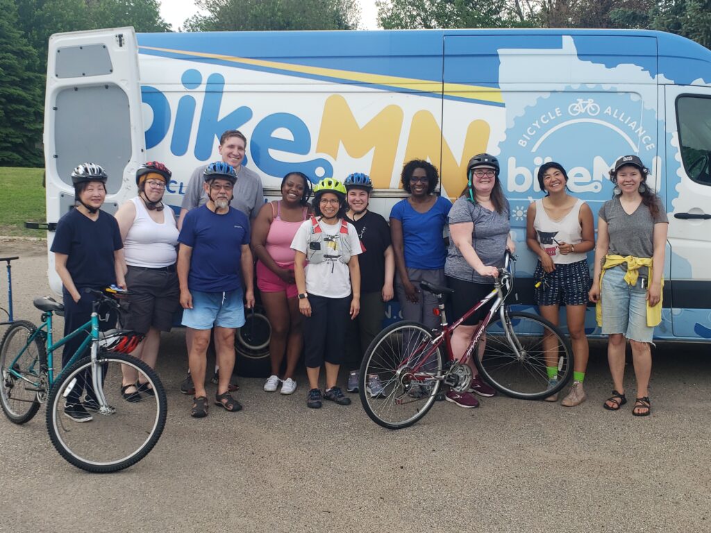 A group of people stand outdoors, some with helmets and some with bikes, in front of the BikeMN van.