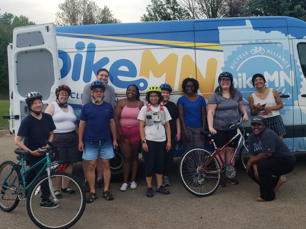 A group of people stand outdoors, some with helmets and some with bikes, in front of the BikeMN van.