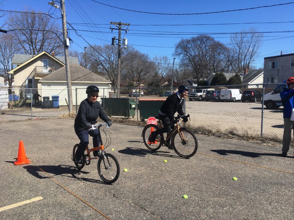 Two Walk! Bike! Fun! participants practice on-bike handling skills outside of the BikeMN headquarters. There are traffic cones to provide direction.