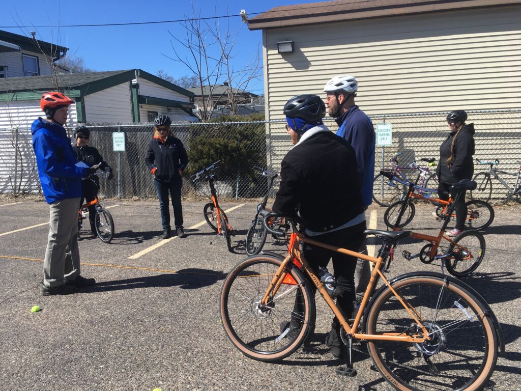 CJ stands in a blue jacket and is surrounded by five instructors who are off of their orange bikes, CJ is providing instructions for the on-bike drills.