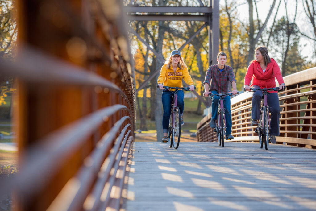Photo of a nature trail bridge at Gooseberry Park in Moorhead, Minnesota during the fall season. Three college-aged people are riding their bicycles across the bridge. One person is wearing a sweatshirt that reads Concordia Cobbers.