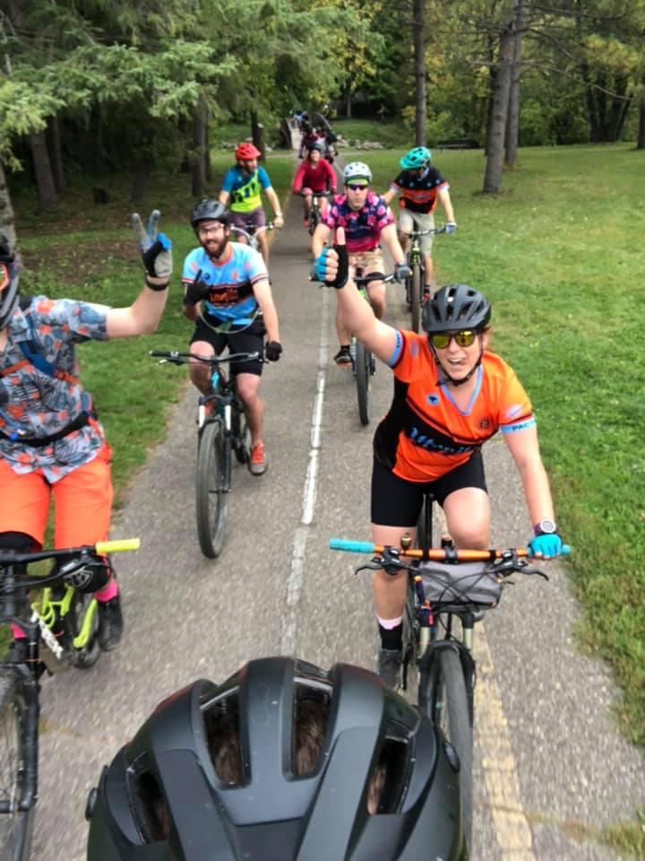 A group of bicycle riders, two abreast, pose for a picture while riding on a bicycle path in a park. The lead rider's helmet shows at the bottom of the picture as they take the picture of the group behind them. One rider flashes a thumbs-up. Another rider is showing the V sign.