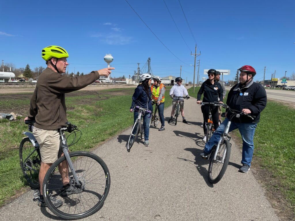 A group of over five people stand with their bikes, facing and listening to the instructor. The Mahnomen water-tower is in the background, and there is a blue sky